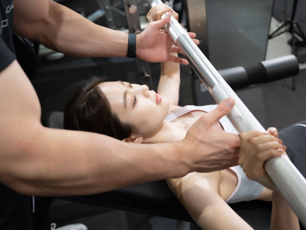 女性が行うベンチプレスを男性トレーナーが補助している